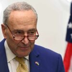 Majority Senate Leader, Chuck Schumer in suit with glasses, American flag background.