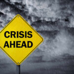 A yellow warning sign that reads 'CRISIS AHEAD' against a stormy sky