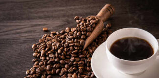 A cup of hot coffee next to spilled coffee beans and a wooden scoop