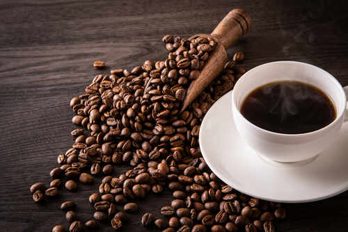 A cup of hot coffee next to spilled coffee beans and a wooden scoop