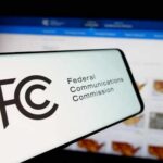 Close-up of a mobile device displaying the FCC logo with a website in the background