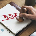 A hand holding a wooden stamp over a document marked 'PROOF'