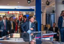 Man seated at news desk with crowd background.