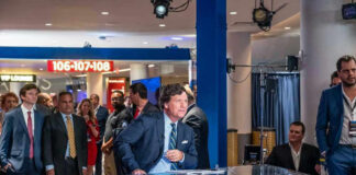 Man seated at news desk with crowd background.