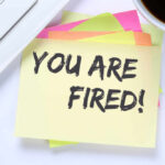 A sticky note with the message 'YOU ARE FIRED' placed on a desk next to a keyboard and a cup of coffee