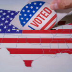 Hand placing a 'VOTED' sticker into a ballot box shaped like the United States