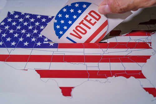 Hand placing a 'VOTED' sticker into a ballot box shaped like the United States