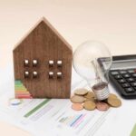 A wooden house model, light bulb, coins, and a calculator on financial documents