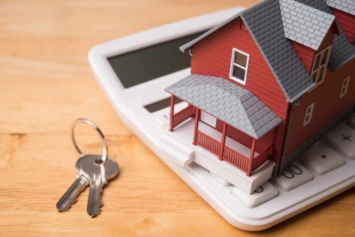 Red,House,Model,On,White,Calculator,And,House,Key,On A miniature red house on a calculator with keys nearby