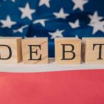 Debt Bubble Swells — What Pops First? Wooden blocks spelling 'DEBT' on a background of the American flag