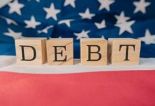 Debt Bubble Swells — What Pops First? Wooden blocks spelling 'DEBT' on a background of the American flag