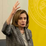 Nancy Pelosi gestures while speaking in front of yellow backdrop.