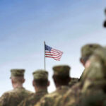 Soldiers in uniform facing American flag outdoors.