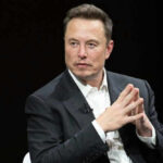 Musk Money Tsunami Hits Midterms Man in suit, hands clasped, black background.
