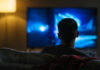 TV Titan Exits – Why Now? Person sitting on a couch watching a television screen in a dimly lit room