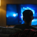 TV Titan Exits – Why Now? Person sitting on a couch watching a television screen in a dimly lit room