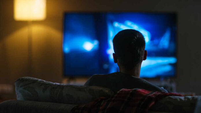 Back,View,Of,A,Man,Sitting,On,A,Couch,Watching Person sitting on a couch watching a television screen in a dimly lit room