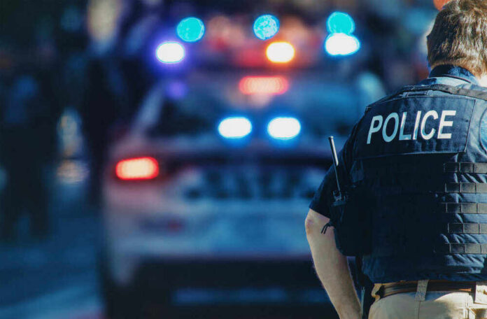 shutterstock_1543591088-1-1 Police officer stands near patrol car with lights flashing.