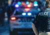 Daybreak Bloodbath Stuns Lib City Police officer stands near patrol car with lights flashing.