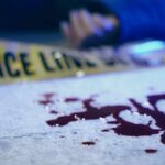 Blood on pavement with police tape and a hand in the background