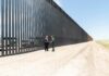 Two people stand by a tall border fence on a dusty road