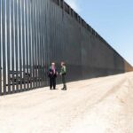 Two people stand by a tall border fence on a dusty road