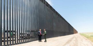 Two people stand by a tall border fence on a dusty road
